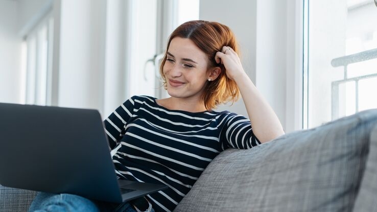 Eine Frau sitzt auf der Couch und lächelt mit Blick in ihren Laptop.